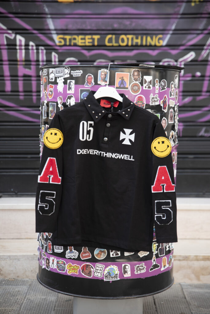 Maglia streetwear nera con dettagli ricamati, numeri, lettere rosse e patch smile gialli su sfondo urbano con adesivi.