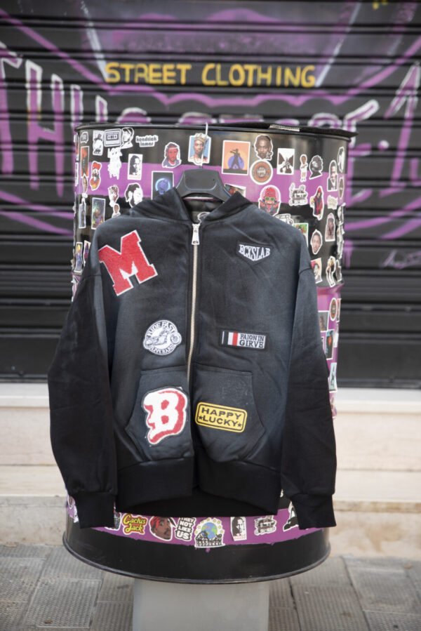 Felpa nera con cappuccio e patch colorate, esposta su sfondo urbano con graffiti per moda streetwear.
