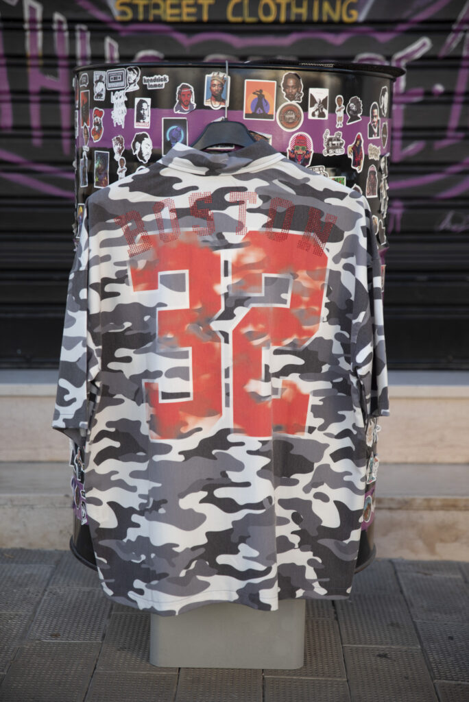Maglia mimetica streetwear con scritta "Boston 32" in rosso, esposta su un manichino all'esterno di un negozio.
