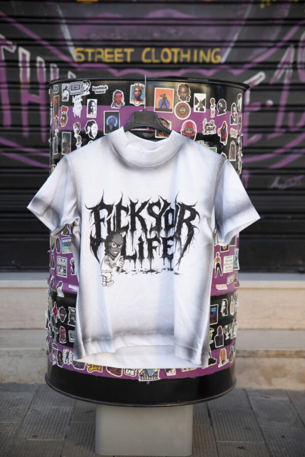 T-shirt bianca con scritta "Fuck Your Life" in stile gotico, esposta su un supporto decorato con adesivi colorati.