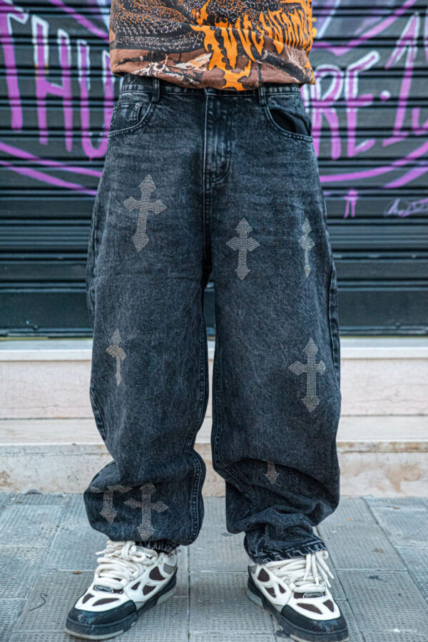 Pantaloni larghi neri con croci ricamate e sneakers bianche, sfondo urbano con graffiti.
