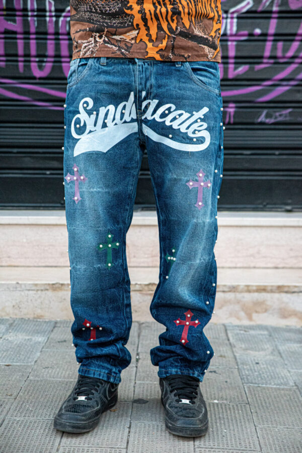 Jeans Straight Fit Sindacate TPWR con croci colorate e scritta bianca, indossati con sneakers nere e maglietta fantasia.