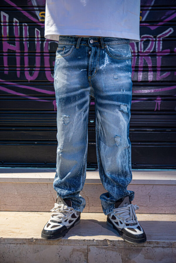Jeans baggy blu lavaggio chiaro con dettagli strappati e sneakers bianche e nere su sfondo urbano.