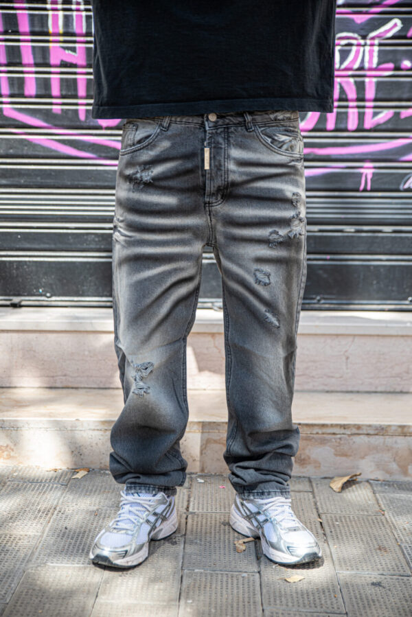 Jeans straight fit grigi con effetto usurato e strappi, abbinati a scarpe da ginnastica bianche su sfondo urbano.