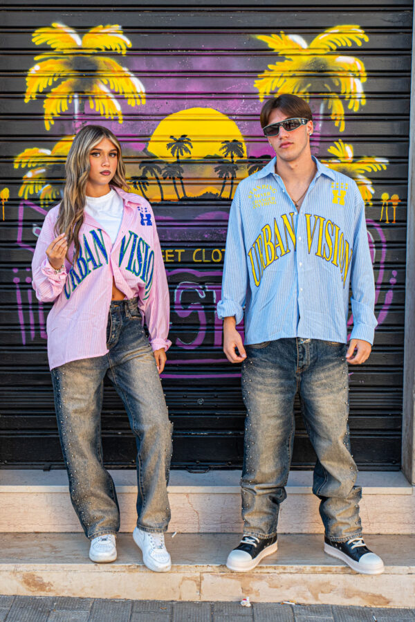 Due persone indossano camicie a righe "Urban Vision" e jeans larghi davanti a un murale tropicale.