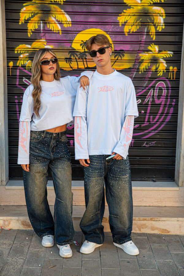Giovani con occhiali da sole indossano maglie bianche e jeans larghi davanti a un murale di palme e tramonto.