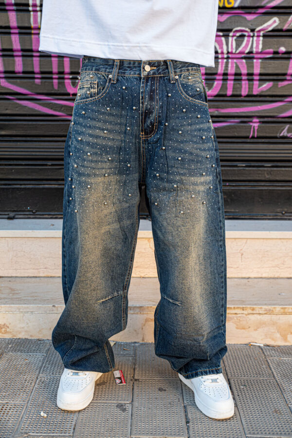 Jeans larghi in denim scuro con perline decorative, abbinati a scarpe bianche sportive, su sfondo urbano con graffiti.