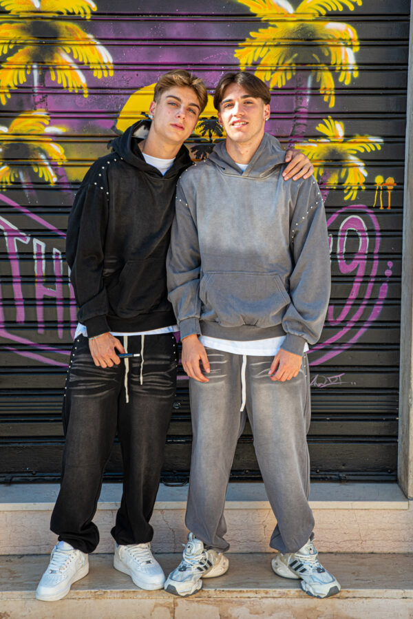 Due ragazzi indossano felpe oversize nere e grigie con pantaloni coordinati davanti a un murale colorato di palme.