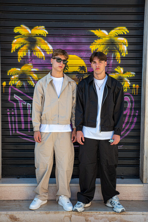 Due giovani uomini in abiti casual coordinati beige e nero, davanti a un murale tropicale colorato.