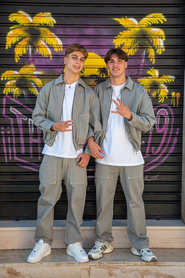 Due ragazzi in abbigliamento casual grigio con t-shirt bianche posano davanti a murale con palme e tramonto.