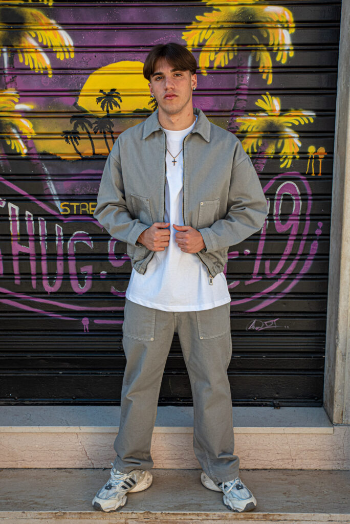 Giovane uomo in abbigliamento casual con giacca e pantaloni grigi davanti a murale colorato con palme e tramonto.