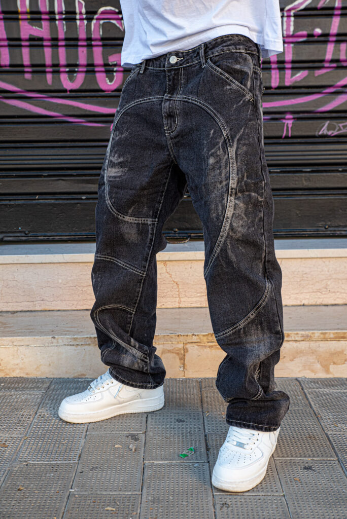 Jeans neri larghi con cuciture decorative indossati con scarpe sportive bianche su sfondo urbano.