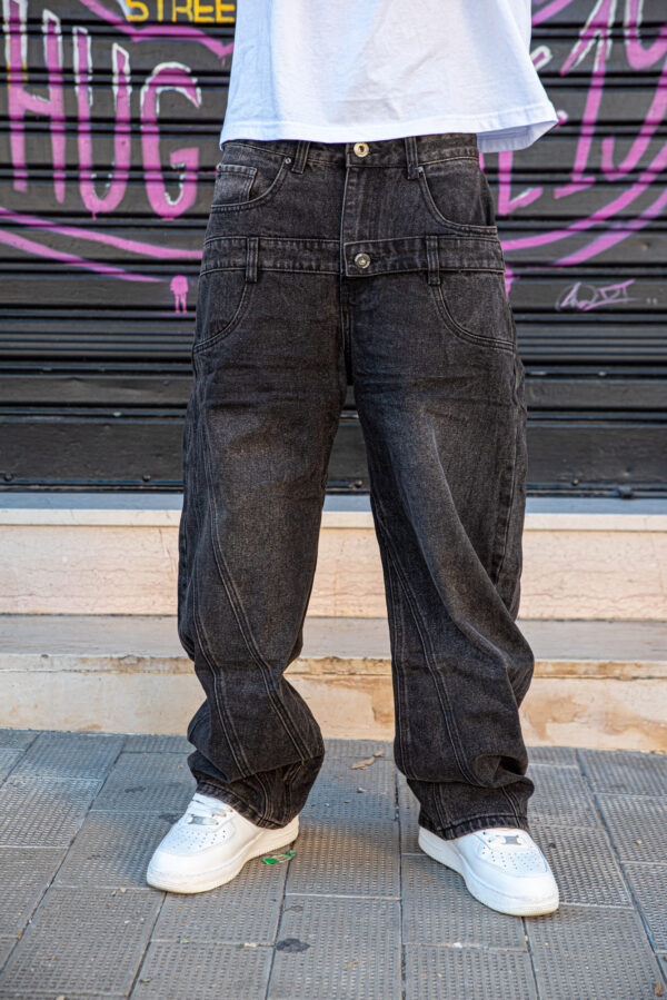 Jeans larghi neri in denim con cuciture vistose, indossati con scarpe da ginnastica bianche su sfondo urbano colorato.