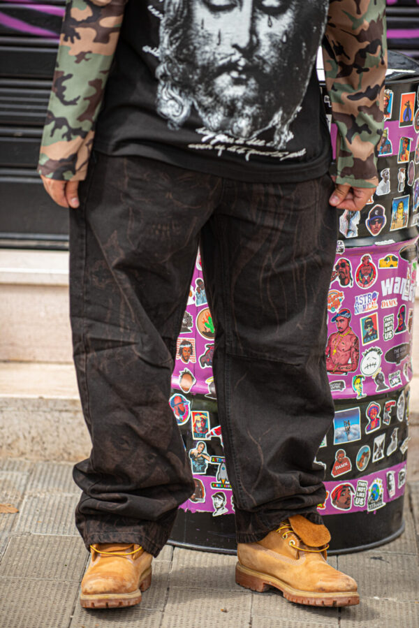 Uomo con pantaloni baggy neri, stivali gialli e maglia nera con stampa artistica, sfondo di adesivi colorati.